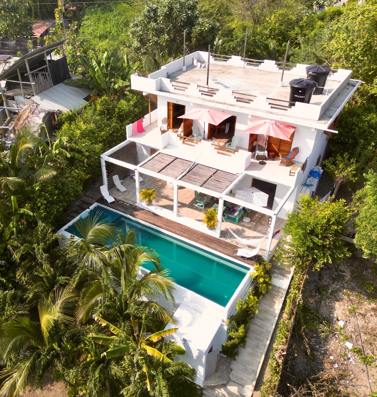 An aerial view of the property highlights a modern building surrounded by lush greenery. A swimming pool is visible at the lower level, with lounge chairs arranged nearby. The rooftop terrace features shaded seating, emphasizing the inviting outdoor spaces for relaxation.