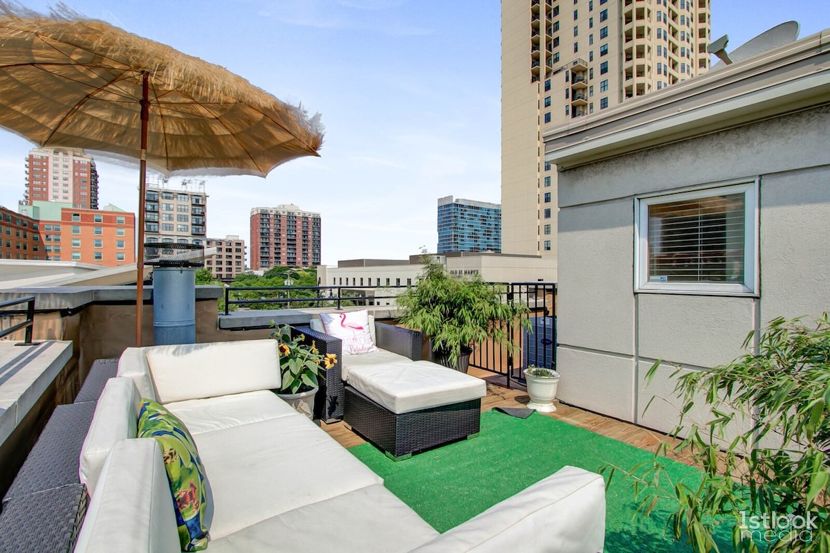 A rooftop terrace is showcased, featuring a comfortable seating area with white lounge furniture and a vibrant green rug. A large umbrella provides shade, while planters with greenery add a touch of nature. The urban skyline is visible in the background.
