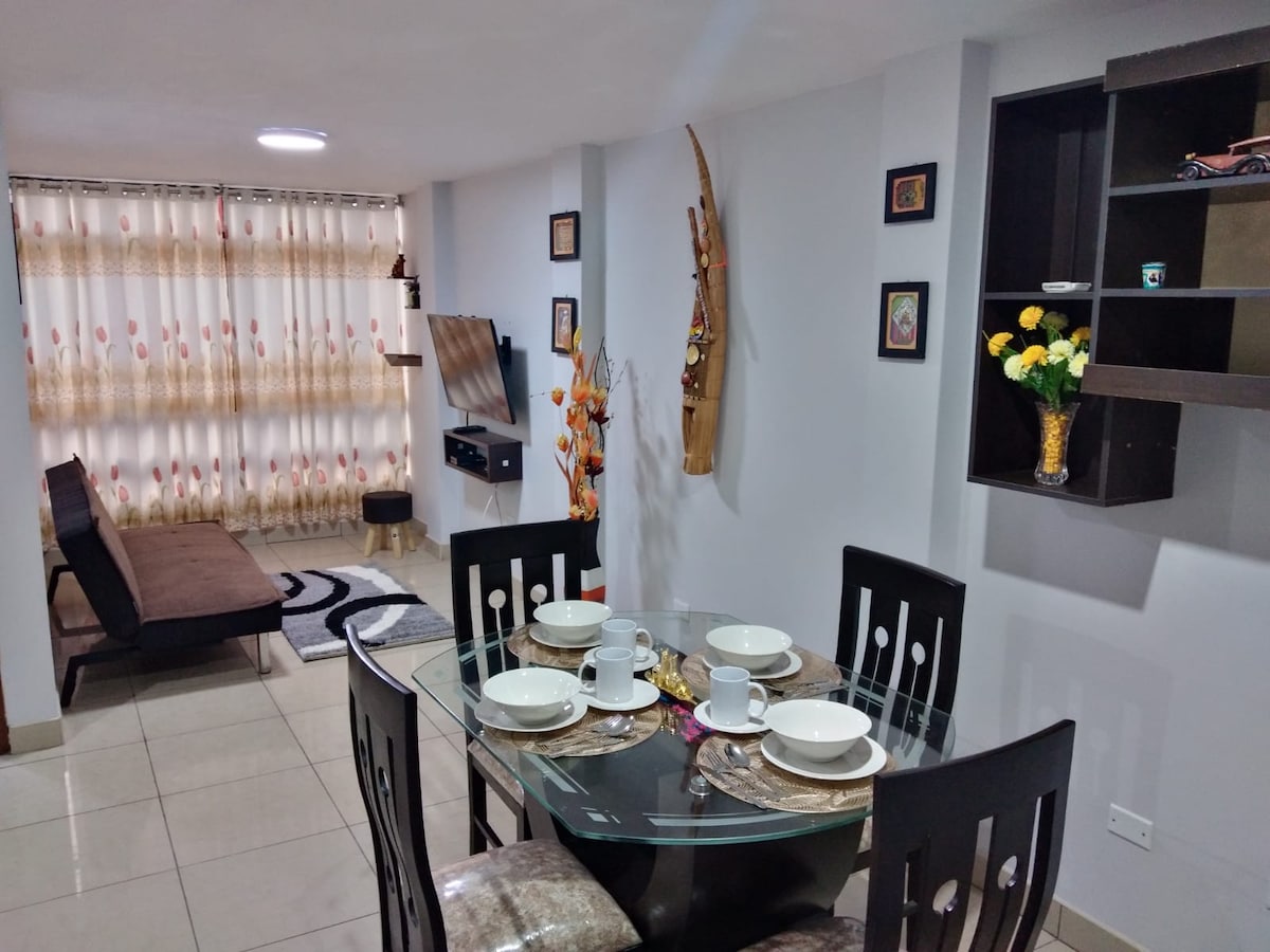 A modern dining area is setup, featuring a glass table with four place settings. A brown sofa is positioned nearby, complemented by patterned curtains allowing natural light to filter in. Decorative wall art can be seen, alongside a flat-screen television mounted on the wall.