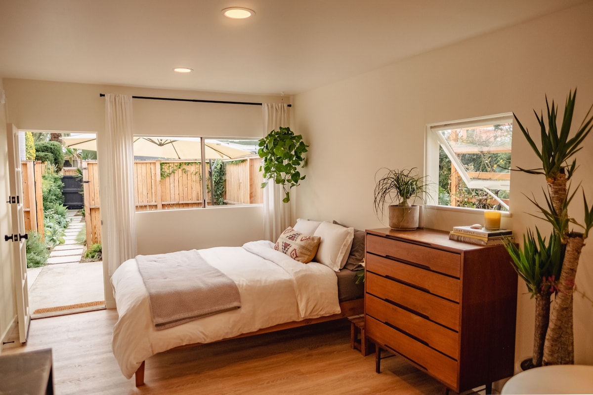 A bright and inviting sleeping area features a spacious bed with neutral bedding. Natural light enters through two windows, illuminating the hardwood floor. A wooden dresser stands beside the bed, complemented by potted plants that add a touch of greenery. An entrance to the outdoor space is visible.