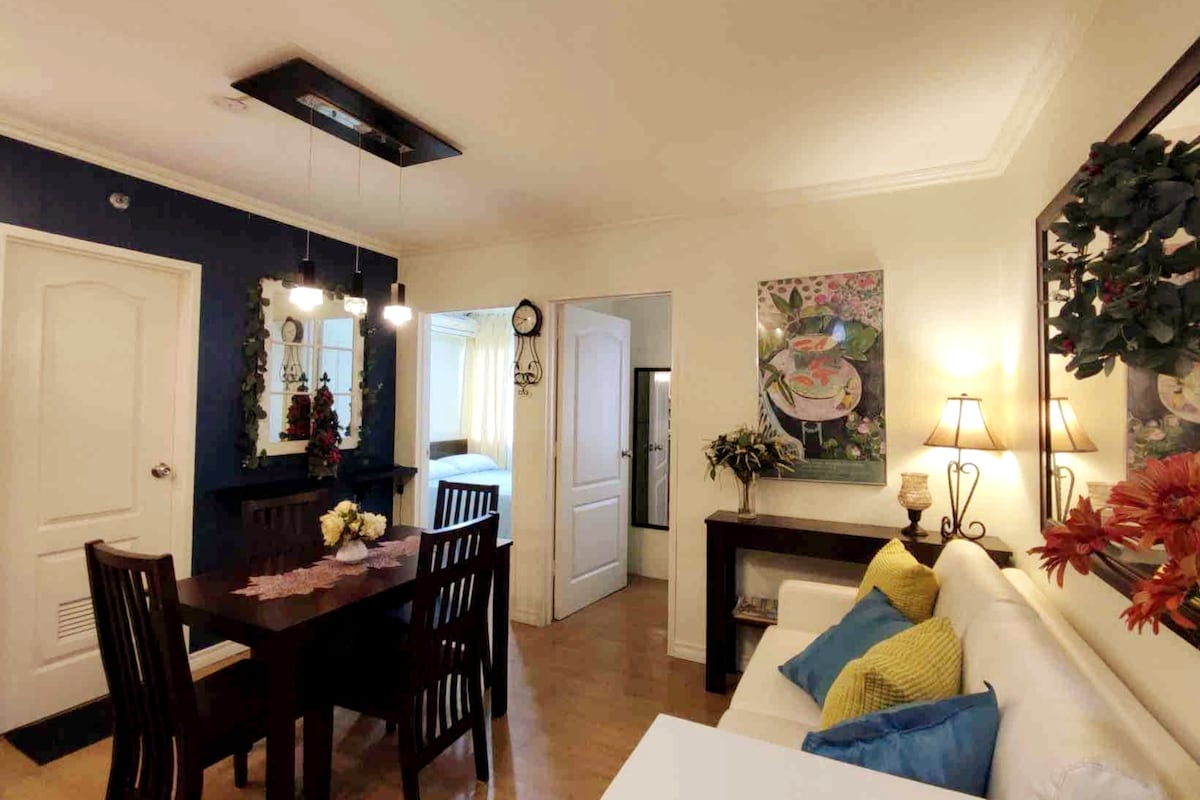 The living area features a cozy seating arrangement with a light-colored sofa and a wooden dining table surrounded by four chairs. Wall art and floral accents add character, while natural light enters through windows. A door leads to the bedrooms, enhancing the unit's spacious layout.
