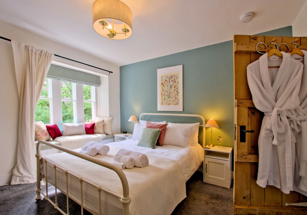 A cozy bedroom features a spacious bed with crisp linens, flanked by bedside tables and soft lighting. A comfortable seating area with decorative pillows complements a large window dressed with light curtains. A wooden door displays plush robes, adding a touch of comfort.