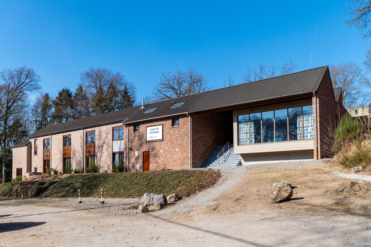 A modern brick building with large windows offers a welcoming view. The structure features a mix of wooden and glass accents, surrounded by a natural landscape. Graveled pathways and landscaped areas lead to the entrance, creating an inviting approach to the accommodation.