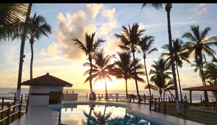 The image depicts a tranquil swimming pool reflecting the sunset, surrounded by tall palm trees. A gazebo is visible near the pool's edge, while lounge chairs offer a space for relaxation. The warm colors of the sky enhance the serene environment.
