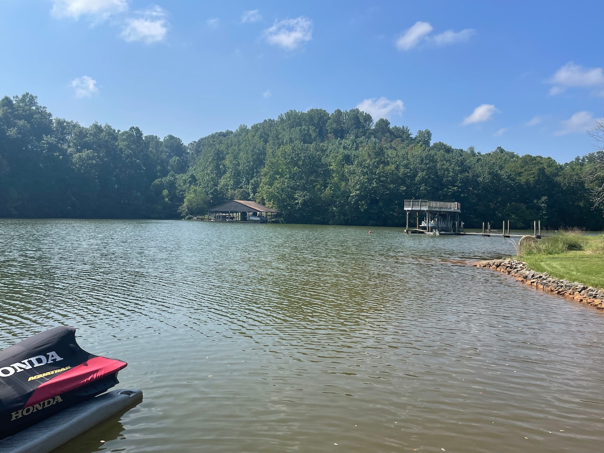 A serene lake scene showcases calm waters reflecting the blue sky and light clouds. Lush greenery lines the shoreline, while a dock is visible across the water, nestled among the trees. In the foreground, a portion of a boat is partially submerged in the water.