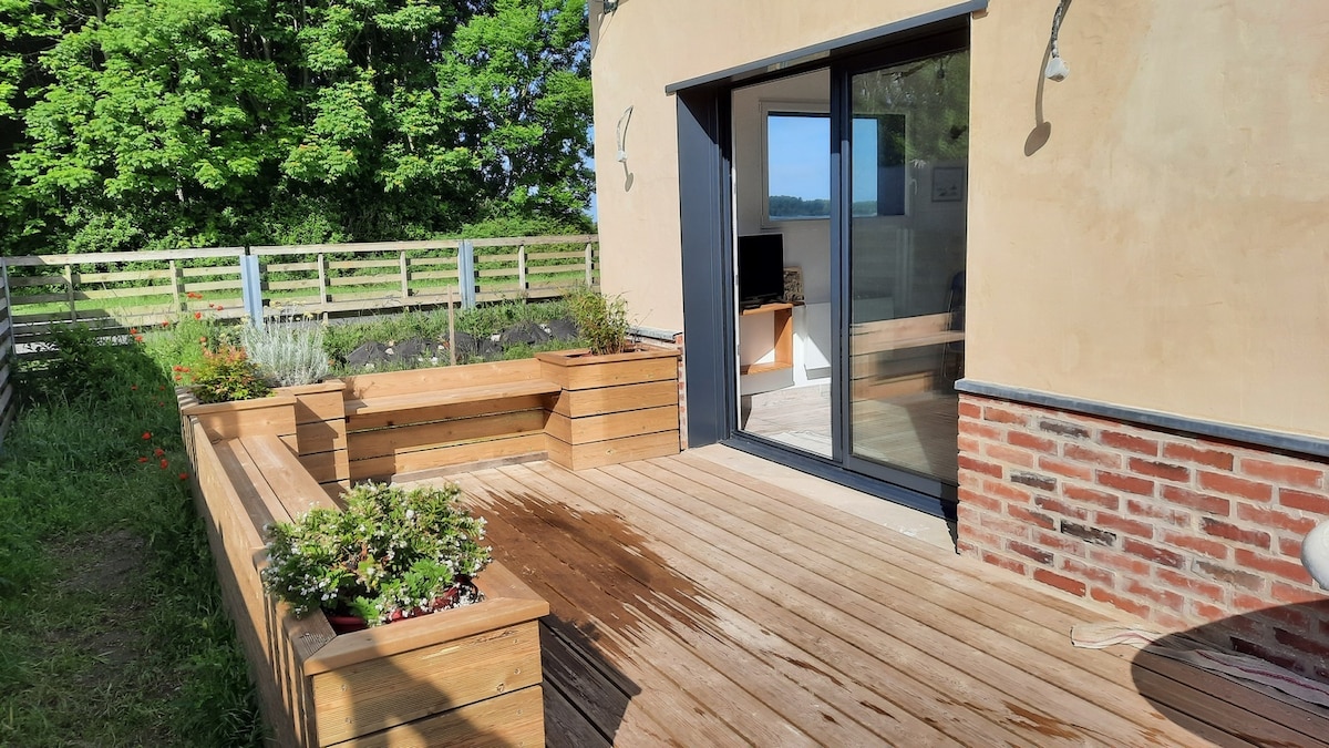 A wooden deck area is featured outside, adorned with planters and surrounded by greenery. Large glass doors open into the living space, offering easy access. Natural light enhances the space, while the simple design blends harmoniously with the outdoor environment.