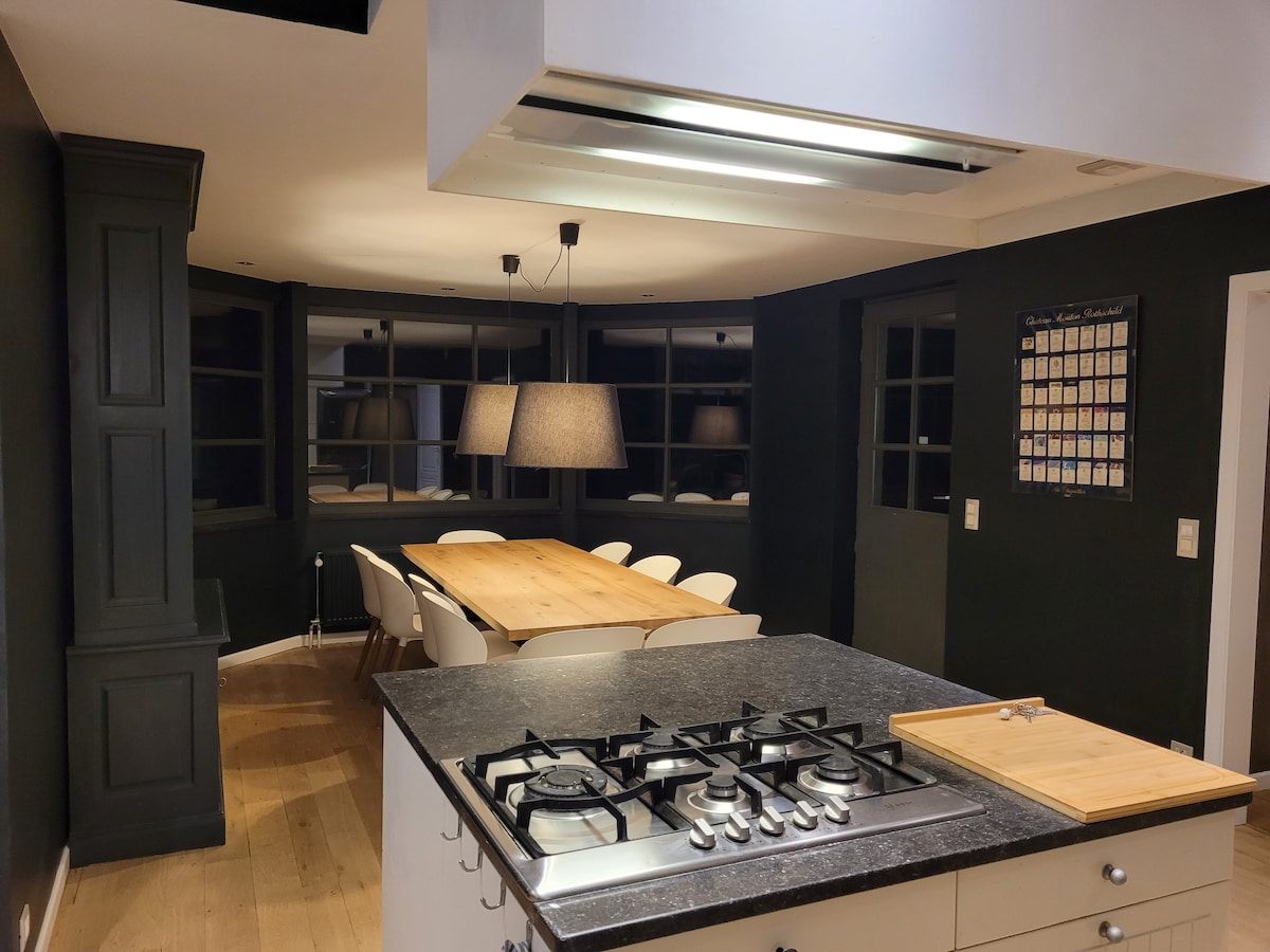 The kitchen features modern appliances, including a gas stove. A dining area is visible, with a large wooden table surrounded by white chairs. Soft lighting creates a welcoming atmosphere, while dark walls and strategically placed windows enhance the room's layout.