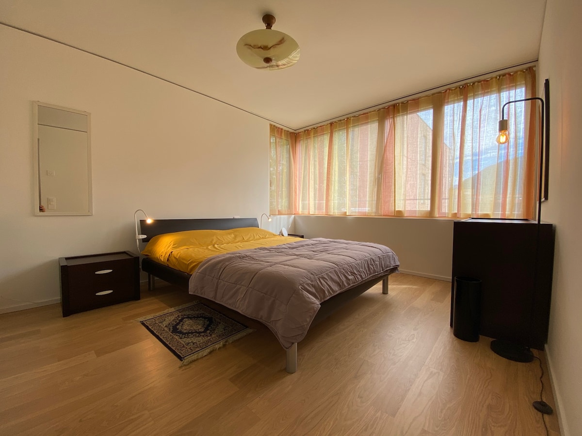 A spacious bedroom features a large bed with a yellow comforter, accompanied by bedside tables. Tall windows dressed in light, flowing curtains allow natural light to fill the room. A sleek lamp and a dark cabinet add functional elements to the minimalist design.