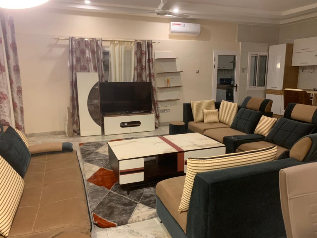 A spacious sitting room features a large, marble-topped coffee table at its center, surrounded by comfortable sofas. A flat-screen television is mounted against the wall, complemented by modern storage units. Large windows are adorned with patterned drapes, allowing natural light to fill the space.