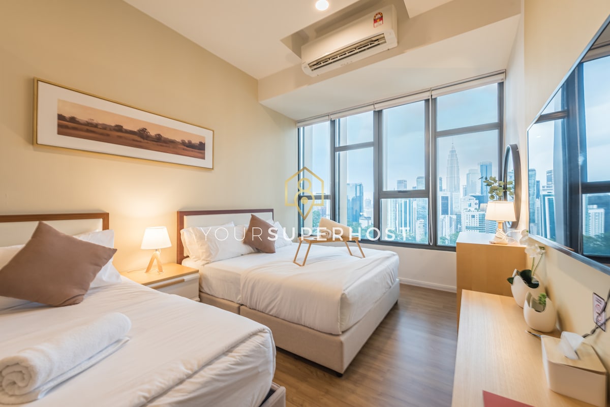 The room features two spacious beds, positioned near large windows that offer views of the city skyline. Soft lighting from bedside lamps complements the neutral wall tones, and a wooden console is visible, enhancing the overall layout.