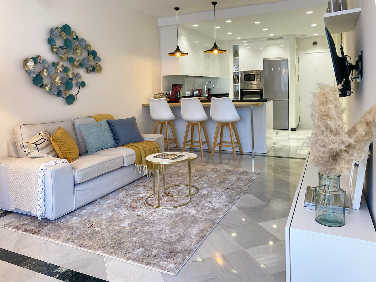 A modern living area features a light-colored sofa adorned with cushions, complemented by a decorative rug on polished marble flooring. Two pendant lights hang above a breakfast bar with three stools, while a TV is mounted on the wall opposite a minimalist entertainment unit.