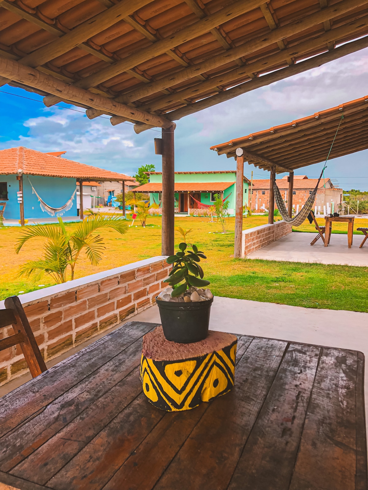 An outdoor seating area features a wooden table with a circular, painted base. A potted plant is positioned at the center. In the background, colorful buildings and hammocks can be seen, surrounded by lush green grass under a partly cloudy sky.
