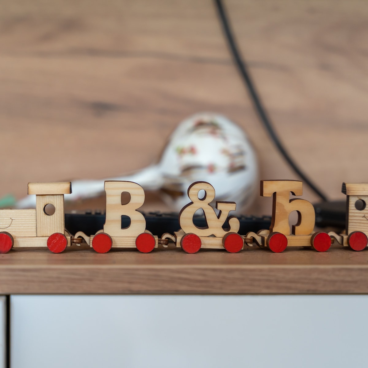 A wooden toy train is displayed, featuring lettered compartments spelling out 'B & B.' The train is mounted on a wooden surface, complemented by a floral ceramic piece blurred in the background, adding a playful touch to the space.