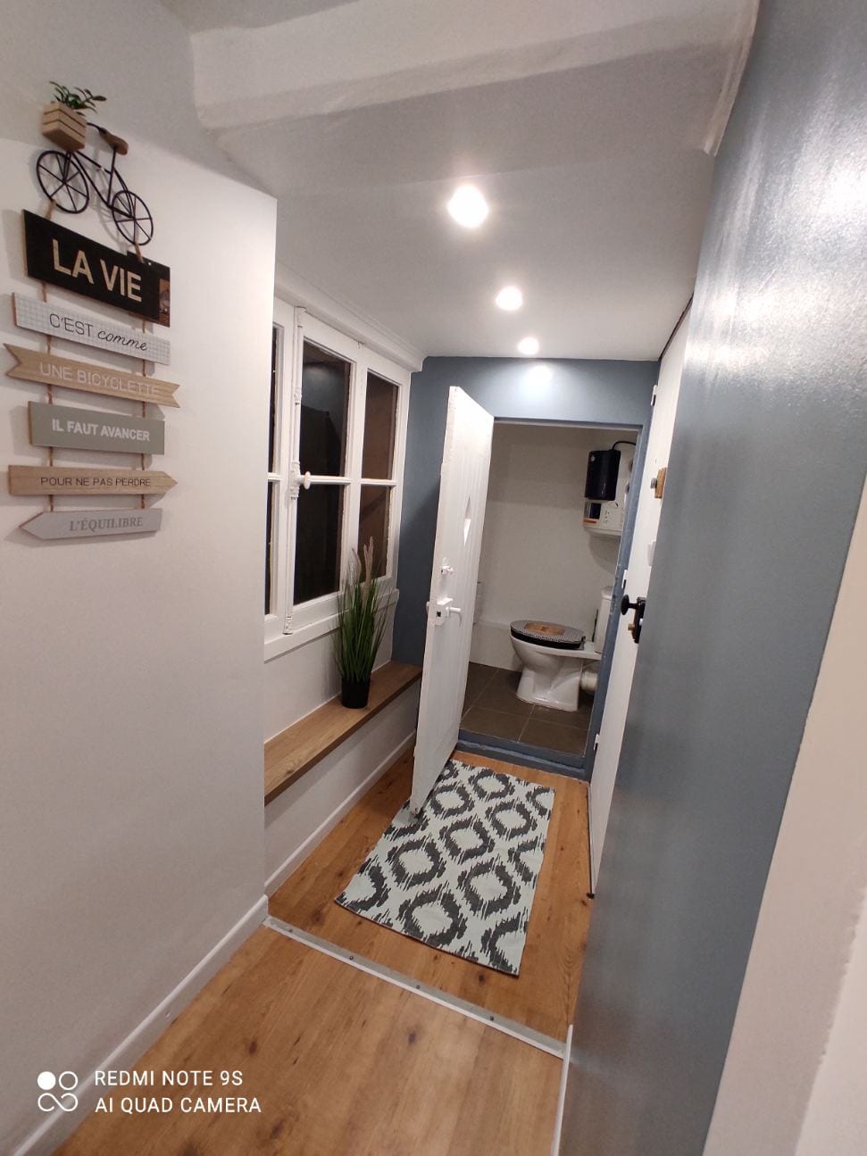 A hallway is featured with a light wood floor and soft blue walls. A small bench is positioned beneath a window with white frames. A decorative sign hangs on the wall, and a rug with a geometric pattern lays on the floor, leading to an open door that reveals the bathroom.