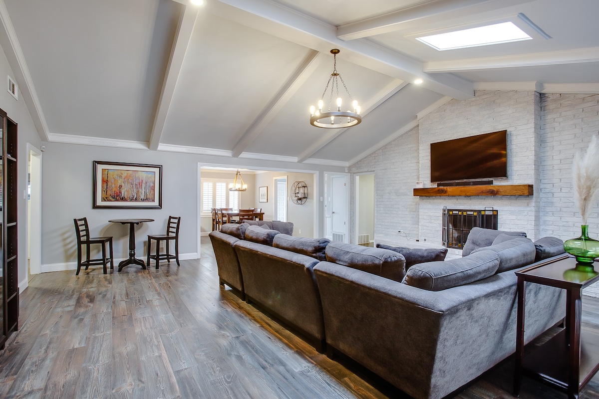 An open living area features a large, L-shaped sofa providing ample seating. Natural light is welcomed through a skylight, highlighting the exposed beams on the ceiling. A small dining table with two chairs and a fireplace with a mounted television enhance the cozy ambiance.