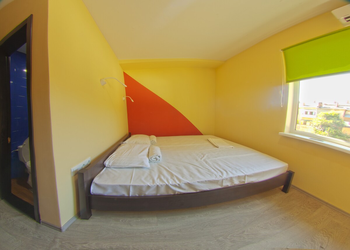 A brightly colored room features a spacious bed with white linens. A vibrant orange accent wall complements the yellow walls. Natural light enters through a window covered with a green roller shade, illuminating the floor. A small wall lamp is positioned above the bed.
