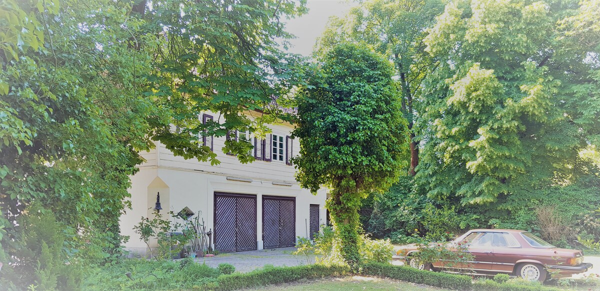 A historic building is nestled among lush greenery, featuring overhead shutters and double wooden doors. A vintage car is parked nearby, complementing the tranquil outdoor setting. The natural surroundings create a serene atmosphere, enhancing the visual appeal of the property.