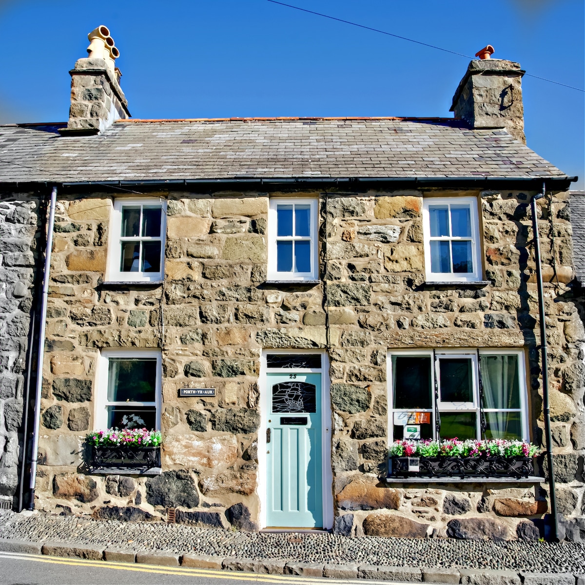 The stone facade of the cottage showcases a charming historical character. Two large windows with flower boxes are displayed on either side of the door, and a light blue front door is centered beneath a decorative arch. The rooftop includes traditional chimney stacks.