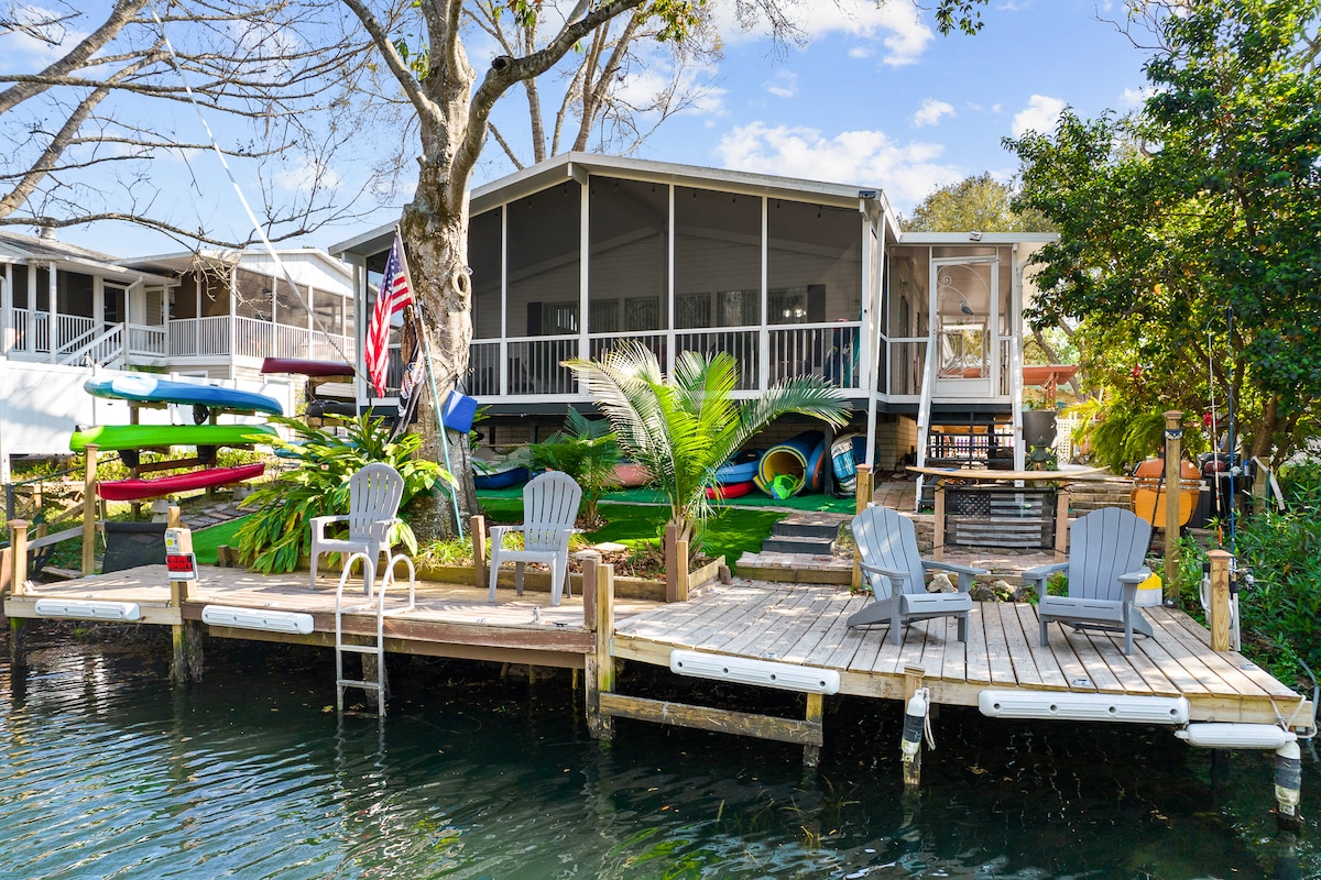 A well-maintained wooden dock is positioned by the canal, featuring several lounge chairs and a ladder for easy access to the water. Colorful kayaks are secured alongside, with lush greenery surrounding the area, and the inviting home is visible in the background.