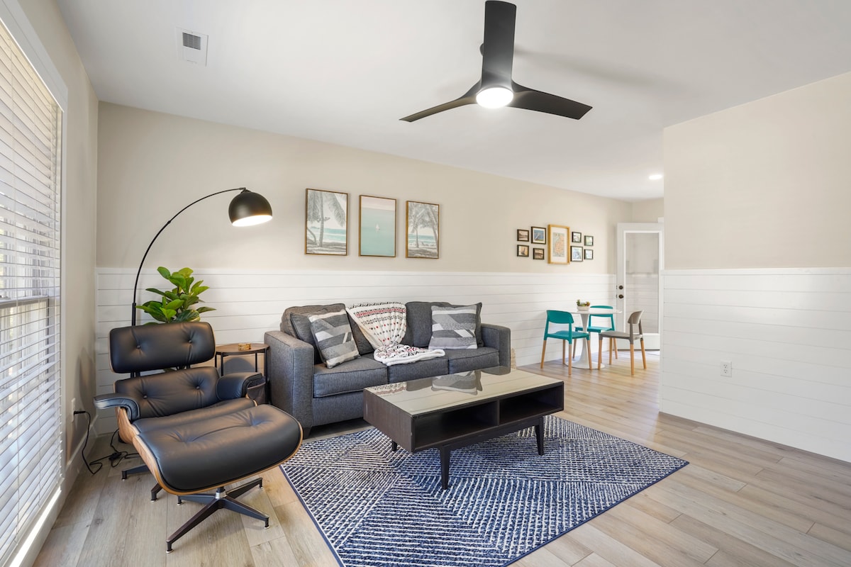 The living room features a modern design with a spacious gray sofa and an iconic Eames leather lounge chair alongside a geometric area rug. Large windows with blinds allow natural light to fill the space, which is accented by a decorative plant and contemporary artwork on the walls.