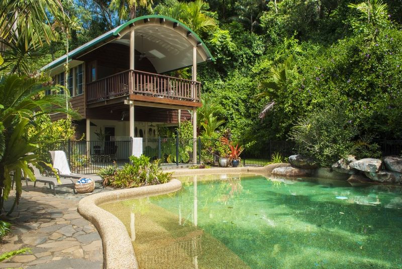 A natural swimming pool is surrounded by lush greenery and tropical plants. A two-story wooden structure features an inviting balcony with railings. Sun loungers are placed beside the pool, offering a serene spot to relax in the secluded setting.