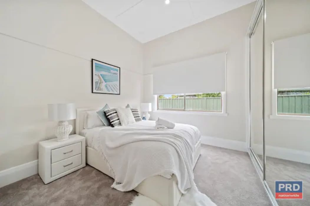The bedroom features a spacious bed adorned with neutral linens and decorative pillows. Bedside tables with lamps are placed on either side. A large mirror wardrobe reflects natural light, while a window provides views of the outdoors, enhancing the bright and airy atmosphere.