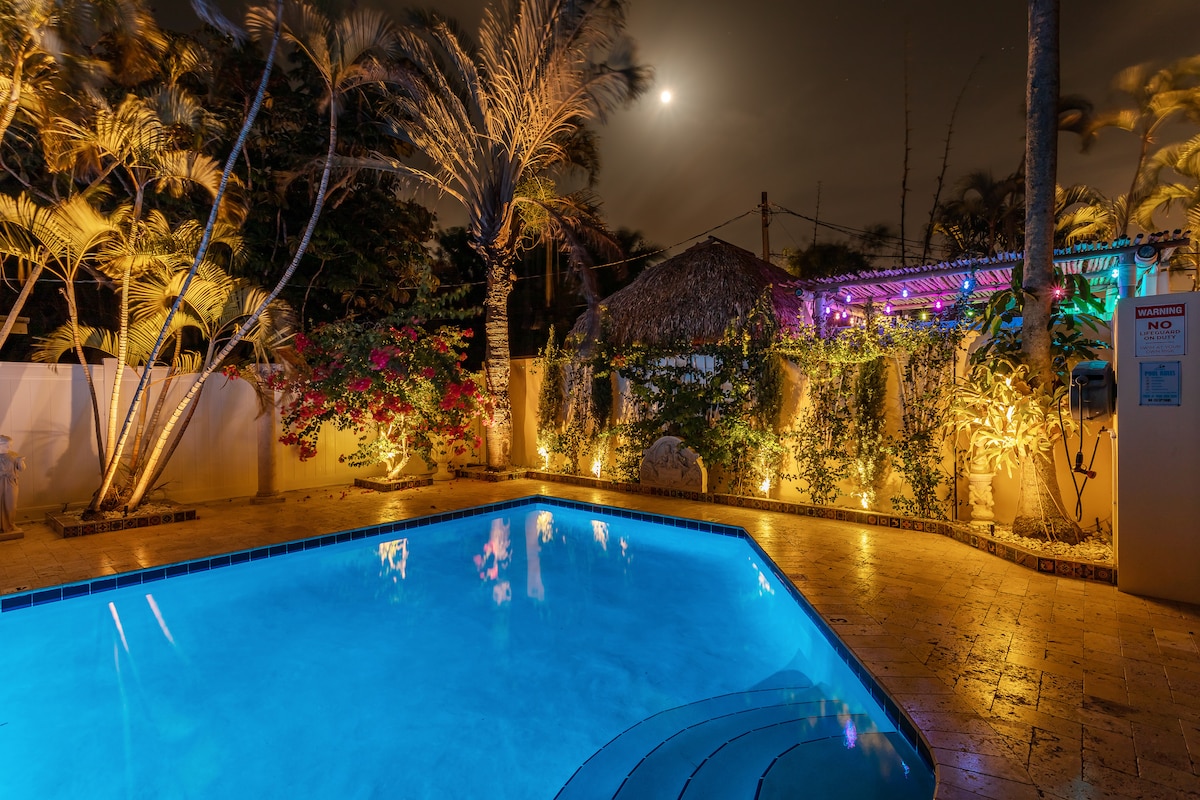 The evening scene showcases a luminous pool illuminated in calming blue, surrounded by tropical plants and vibrant flowers. Soft lighting highlights the lush landscaping and thatched tiki hut in the background, creating a serene atmosphere under a moonlit sky.