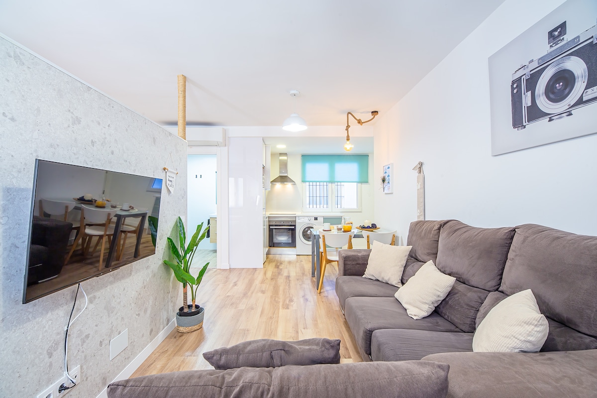 A spacious living area features a comfortable gray sofa with decorative cushions, facing a wall-mounted television. Natural light pours through the window, illuminating the modern kitchen with stainless steel appliances in the background. A dining table with chairs is positioned nearby, complemented by a decorative plant.