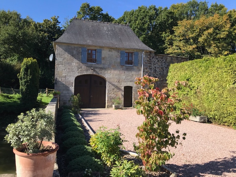 A historic stone building with a steeply pitched roof is set amidst greenery. The entrance features large wooden double doors, while a gravel pathway leads to the front. Flowering plants and neatly trimmed hedges create a welcoming outdoor space.