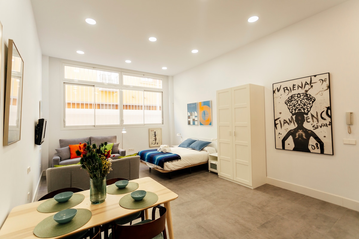 An open-plan loft features a spacious living area with a comfortable sofa and a plush double bed. A dining table set for four, adorned with a vase of flowers, sits near a large window. Contemporary art pieces decorate the walls, enhancing the modern atmosphere.
