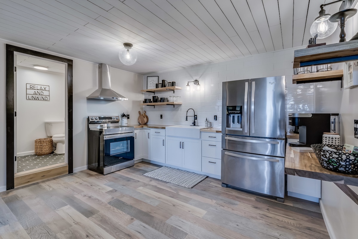 A modern kitchen features stainless steel appliances, including an oven and refrigerator. Open shelving displays decorative items, while white cabinets provide ample storage. The space is complemented by a striped rug and warm wooden accents, enhancing functionality and style in the kitchen area.
