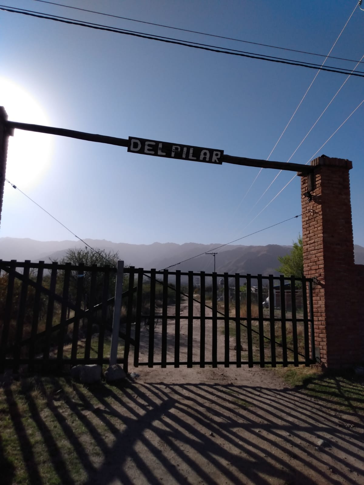 A wooden gate with the name 'DEL PILAR' is prominently displayed at the entrance of a private neighborhood. The sun casts soft shadows on the ground, and the silhouette of mountains can be seen in the background, creating a serene atmosphere.
