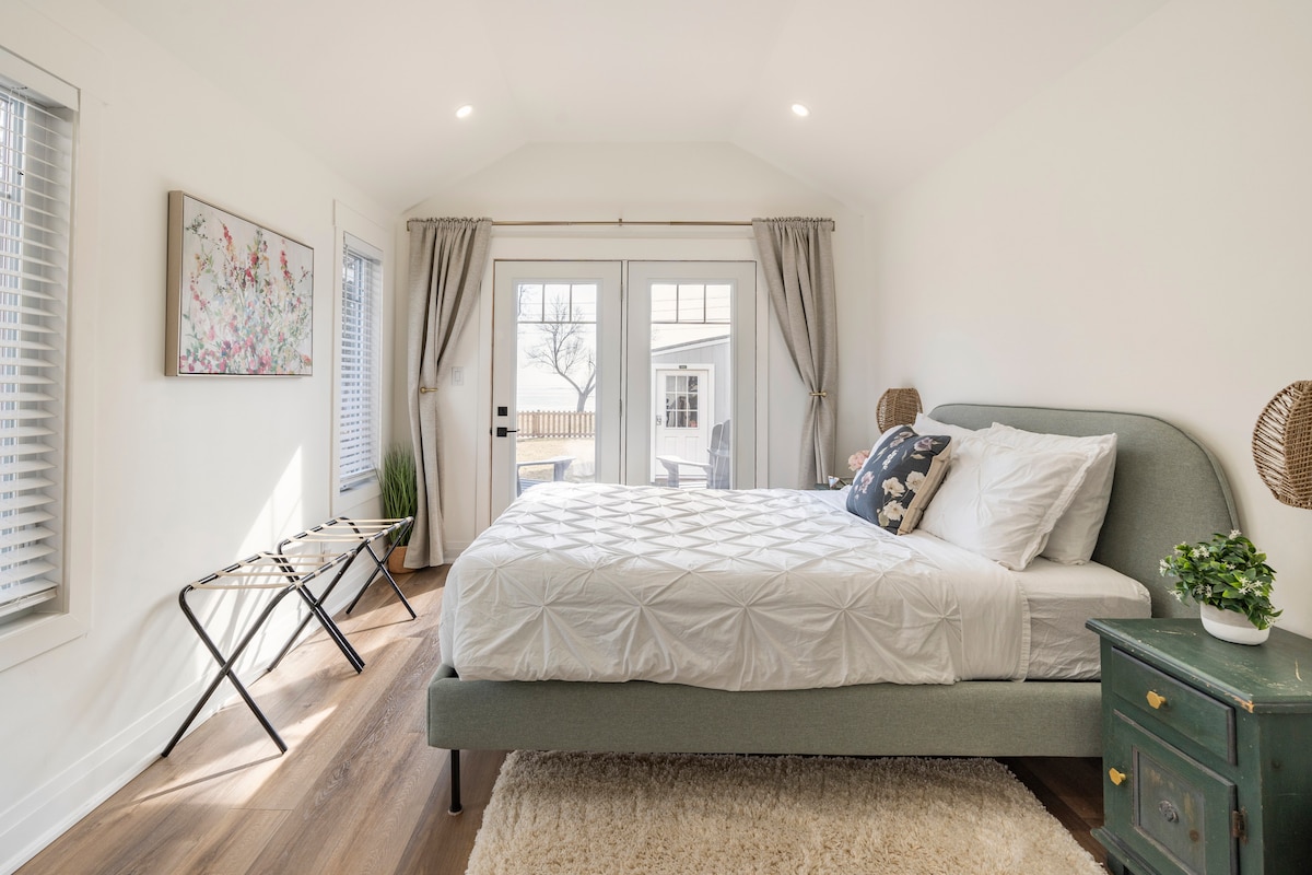 A well-lit bedroom features a spacious bed with a light grey upholstered frame. Double doors open to a view of the outdoors, while two folding chairs are positioned nearby. Simple decor includes a small green plant and artwork on the wall.