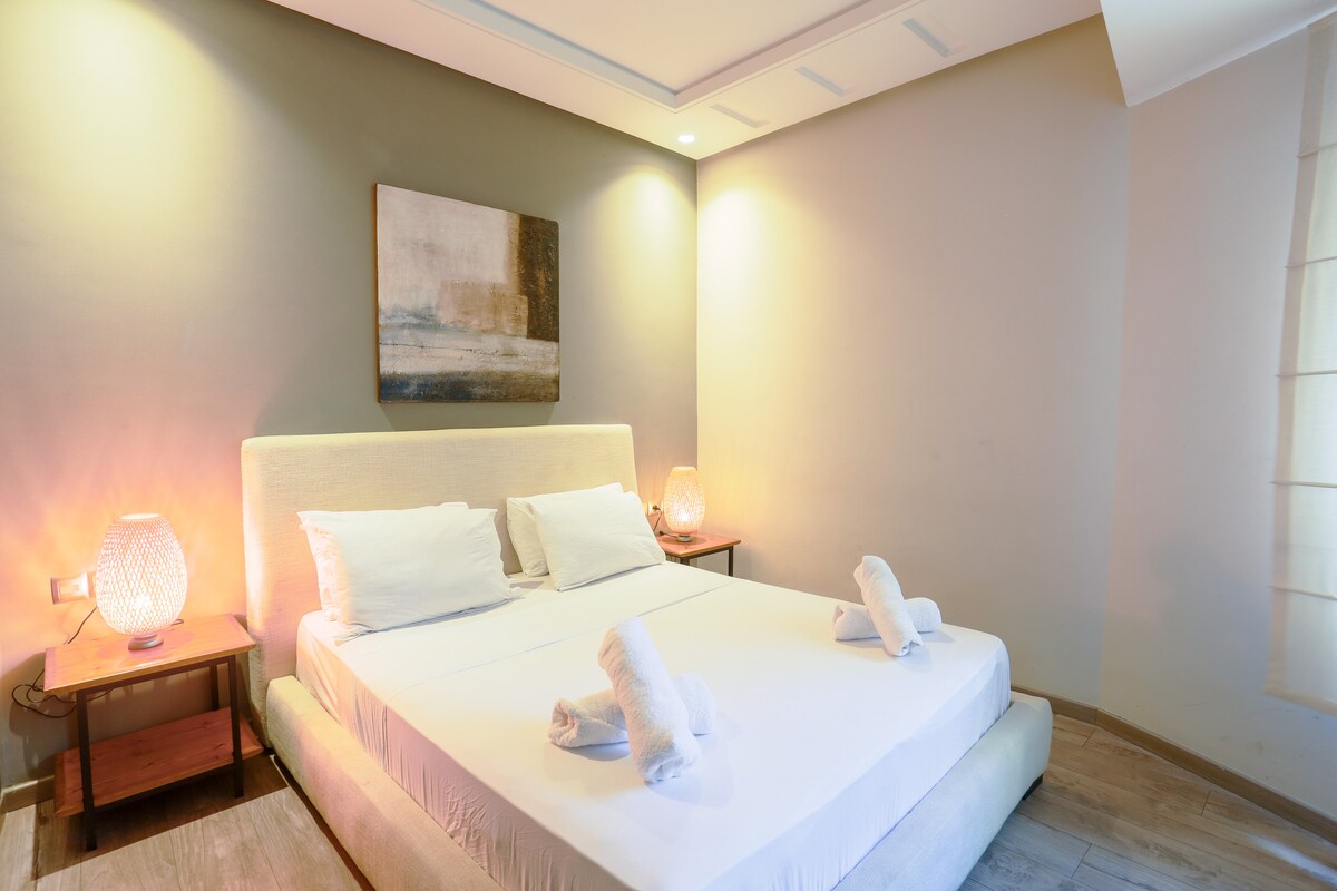 The cozy bedroom features a spacious bed adorned with soft linens. Two bedside tables hold warm lamps that softly illuminate the room. A piece of wall art adds a touch of elegance, while light filtering through the window enhances the overall brightness.