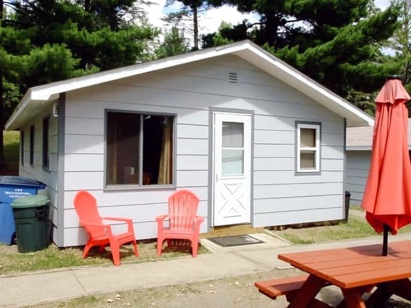 The cabin exterior features a light grey façade with large windows, allowing for natural light. Two red Adirondack chairs are positioned on the porch area, along with a bright umbrella nearby. The entrance showcases a white door framed by simple steps.
