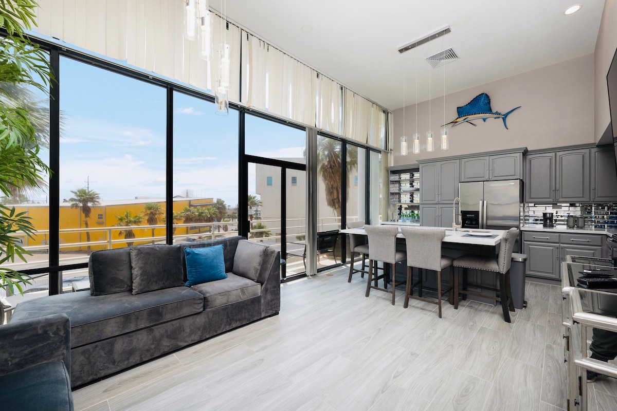 A modern living area features large windows allowing natural light in, with views of the surroundings. A plush, dark sofa is complemented by a blue accent pillow, and a sleek kitchen island with bar seating is visible in the background.