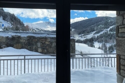 A view from the window reveals a snowy landscape, with rolling hills and distant mountains under a partly cloudy sky. The scene is framed by a sliding glass door and a balcony railing, enhancing the feeling of open space.