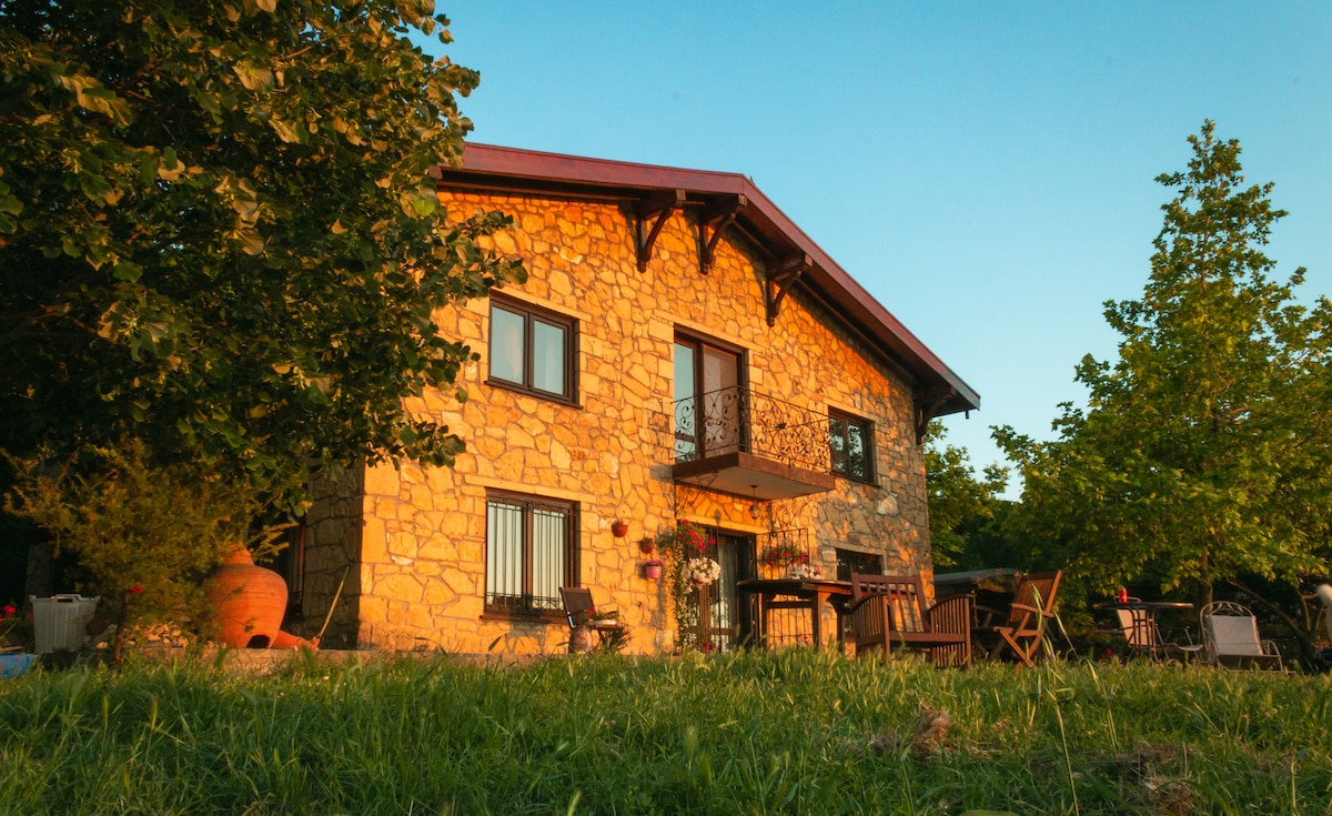 A stone house stands amid lush greenery, features large windows, and a small balcony adorned with decorative ironwork. Various outdoor seating options, including wooden chairs and tables, are positioned on a grass area, inviting relaxation in a natural, tranquil setting.