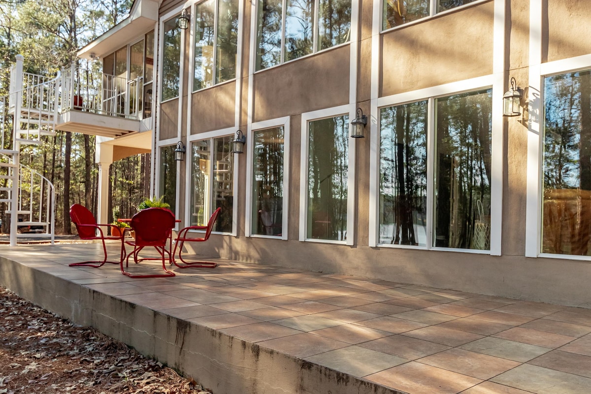 A spacious outdoor patio area features a smooth tiled surface with four red chairs surrounding a table. Large glass windows create an inviting connection to the surrounding nature, and a staircase is visible leading to an upper level, enhancing accessibility and views.