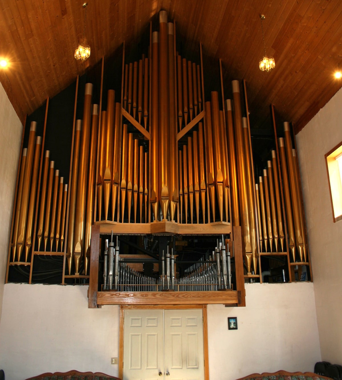 A large pipe organ is prominently displayed on a wooden platform against a backdrop of a vaulted ceiling. The organ is finished in warm tones of gold and wood, with numerous pipes arranged in an impressive formation. Two elegant light fixtures hang above, complementing the serene atmosphere.