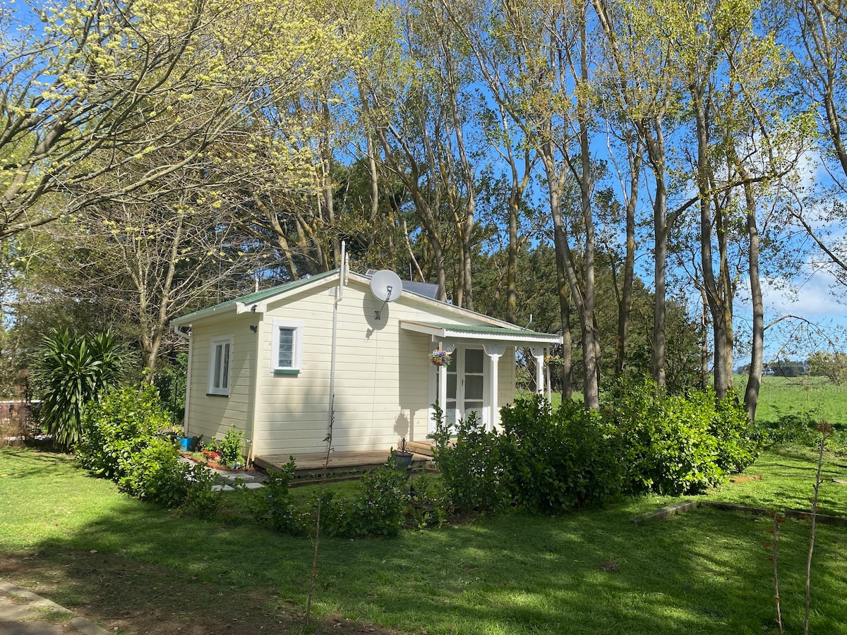 A cottage is nestled among verdant trees and lush greenery, featuring a light exterior and a well-kept garden. Satellite dishes are mounted on the roof, and a welcoming porch is visible. The tranquil setting offers a serene atmosphere, surrounded by expansive farmland.
