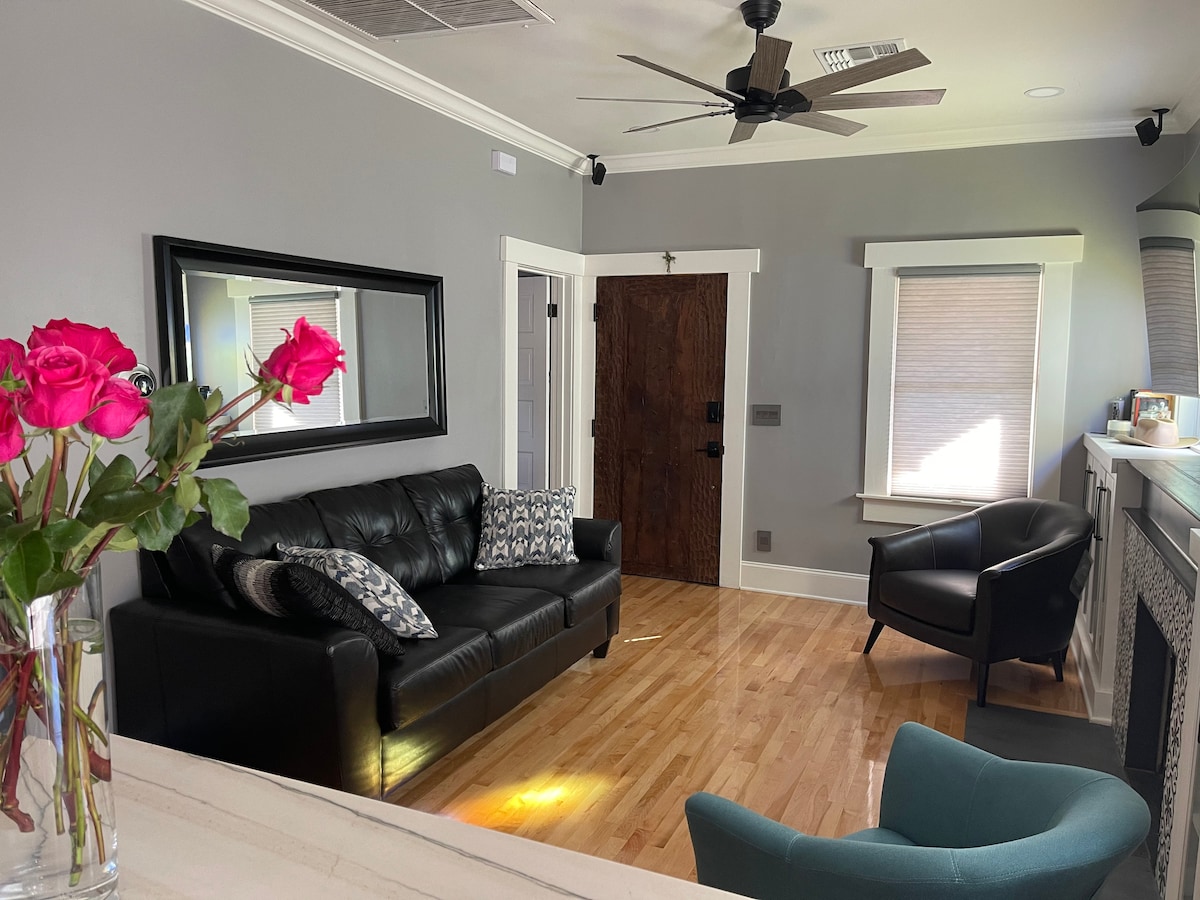 A living area features a black leather sofa adorned with decorative pillows, complemented by a cozy armchair. Natural light filters through the window, highlighting hardwood floors and a wall-mounted mirror. A fireplace adds warmth to the inviting space.