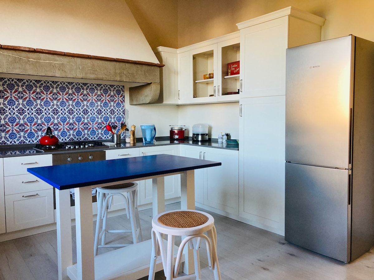 A spacious kitchen features modern appliances, including a stainless steel refrigerator and a gas stove. A large blue island complements white cabinetry, which is highlighted by patterned tiles on the back wall. Two stools are positioned at the island, creating an inviting space for cooking and dining.