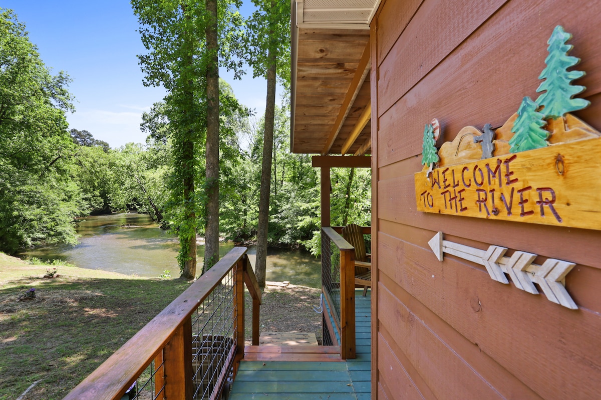 The cabin exterior is framed by lush greenery, with a wooden walkway leading to the entrance. A sign reading 'Welcome to the River' is affixed to the wall. The serene Hiwassee River flows gently in the background, surrounded by trees.