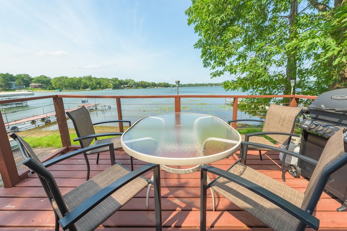 A deck overlooks the lake, featuring a glass-top table surrounded by four chairs. A propane grill is positioned nearby. Lush trees frame the scene, offering shade and enhancing the surrounding natural beauty.