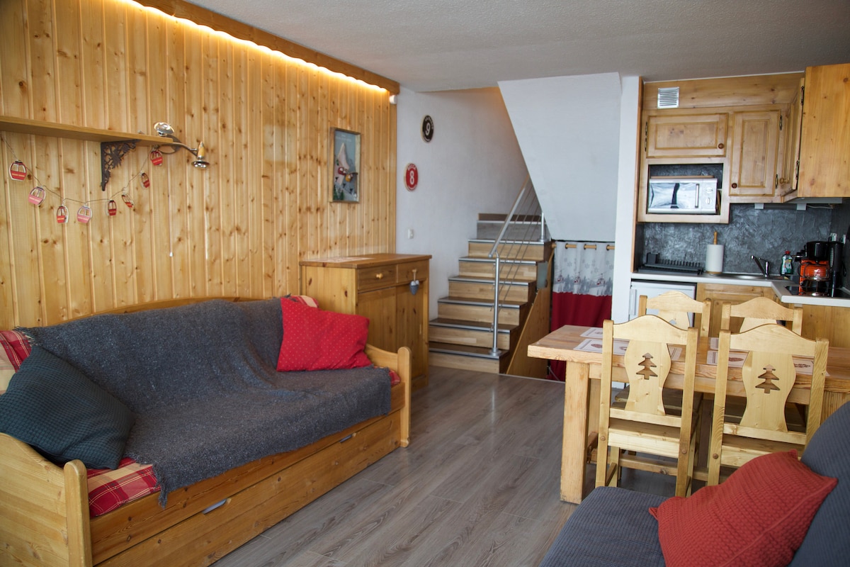 A warm living area features wooden paneling and furnishings, including two sofas with soft throws. A wooden dining table is set with simple chairs. Stairs lead to the upper level, while a kitchenette is visible in the background with modern appliances.