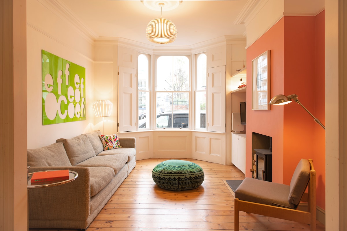 A spacious living area features a large gray sofa, accentuated by a colorful cushion. A green and black pouf adds a unique touch. Natural light floods through bay windows, while an orange accent wall complements the room's bright decor.
