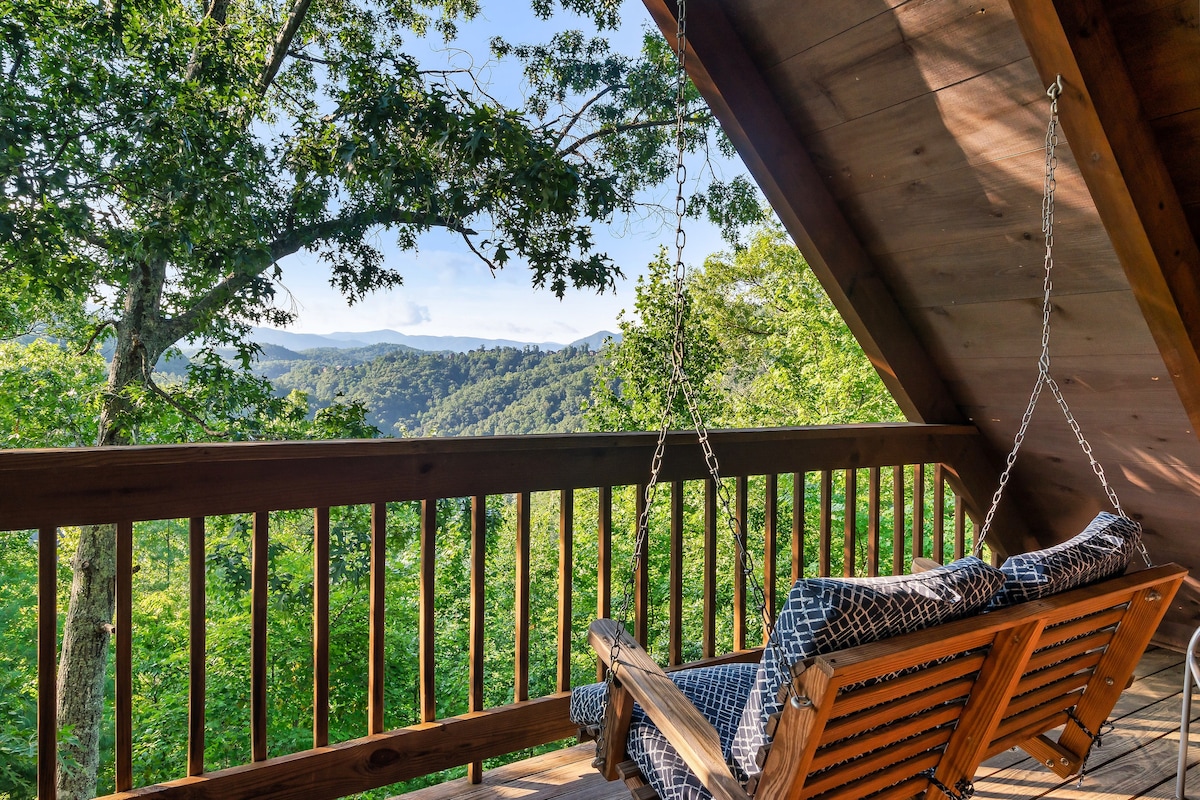 A private upper-level deck offers a porch swing with soft cushions, inviting relaxation amidst the natural surroundings. Lush green trees frame the view, which extends to the distant mountains under a clear sky.