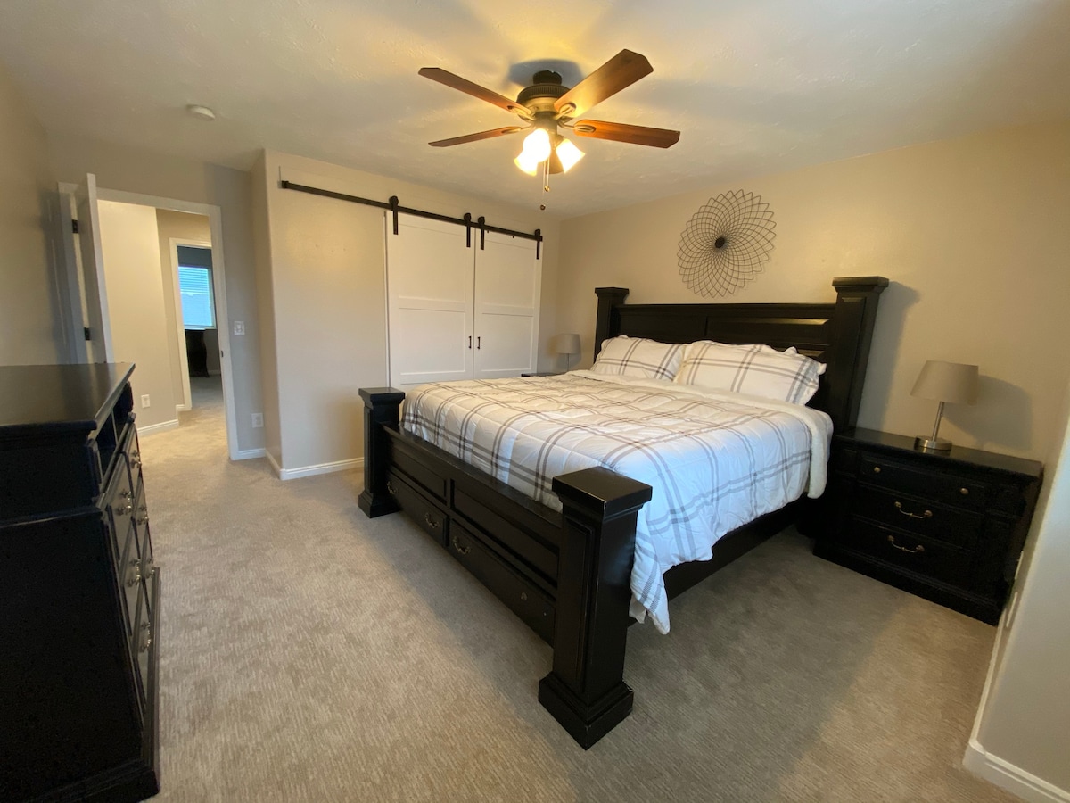 The main bedroom is furnished with a spacious bed featuring a neutral plaid blanket. A ceiling fan provides airflow. Two bedside tables are positioned next to the bed, each with a lamp. A dresser and a sliding barn door add functionality to the inviting space.