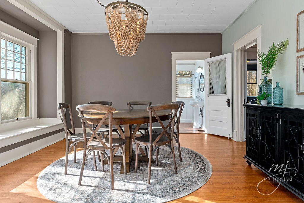 A dining area features a round wooden table surrounded by six chairs, situated on a patterned area rug. Natural light streams in through large windows, illuminating the space. A black cabinet and a chandelier made of beads provide additional texture to the room.